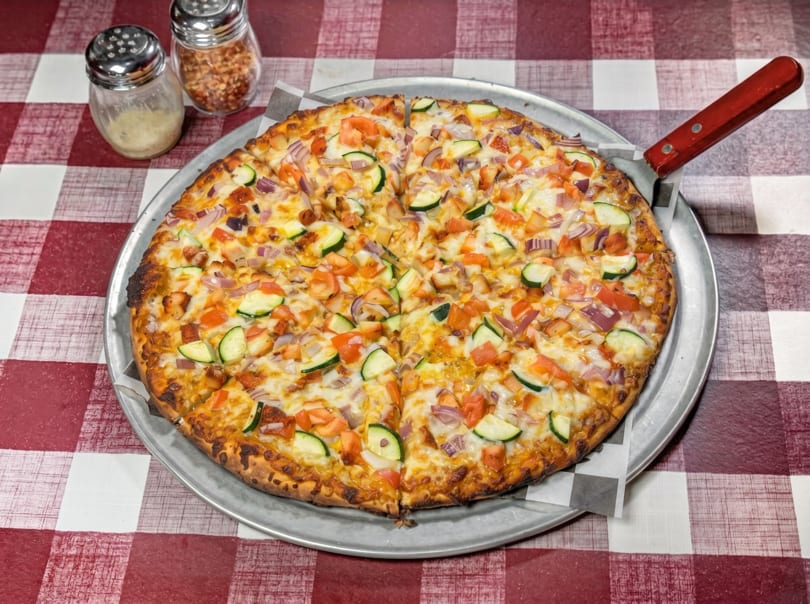 A whole pizza with zucchini, tomatoes, and red onions on a red and white checkered tablecloth.