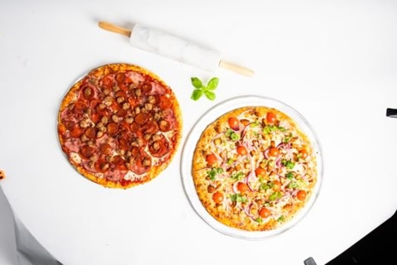 Two pizzas, one meat and one veggie, with a rolling pin and basil on a white surface.