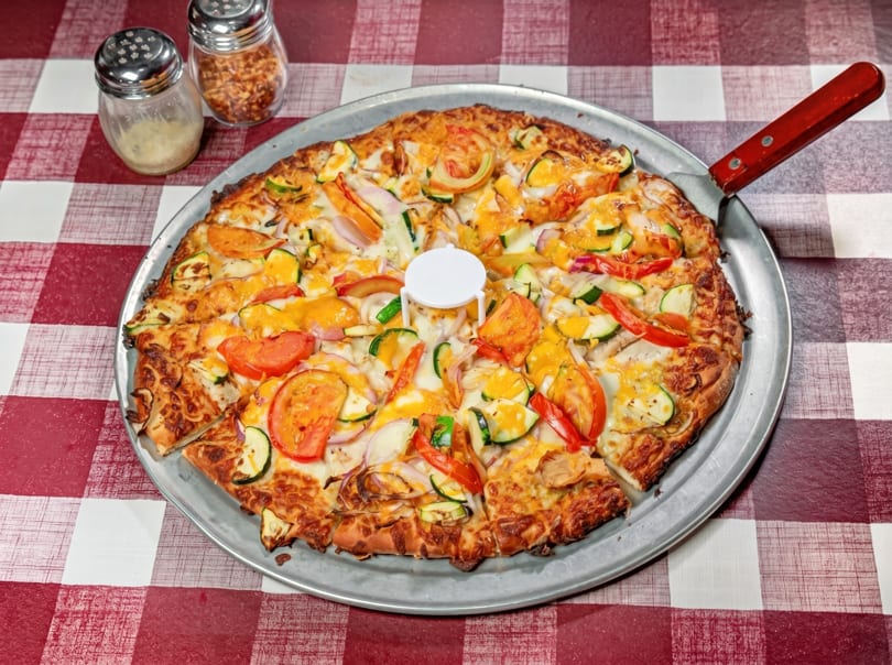 A large vegetable pizza on a metal pan with a red and white checkered tablecloth.