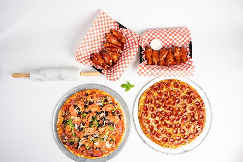 Two pizzas, one pepperoni and one supreme, with two baskets of buffalo wings on a white background.