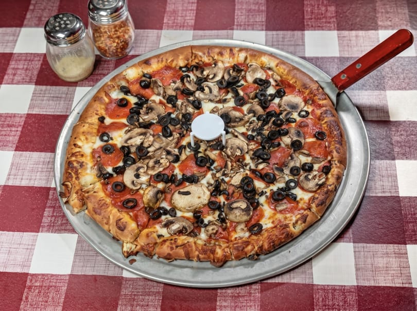 A whole pepperoni, mushroom, and olive pizza on a metal pan with a red and white checkered tablecloth.
