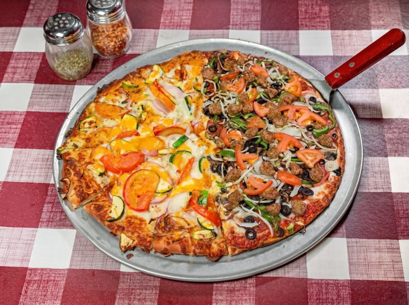 Half veggie, half meatball pizza on a metal pan with a red and white checkered tablecloth.