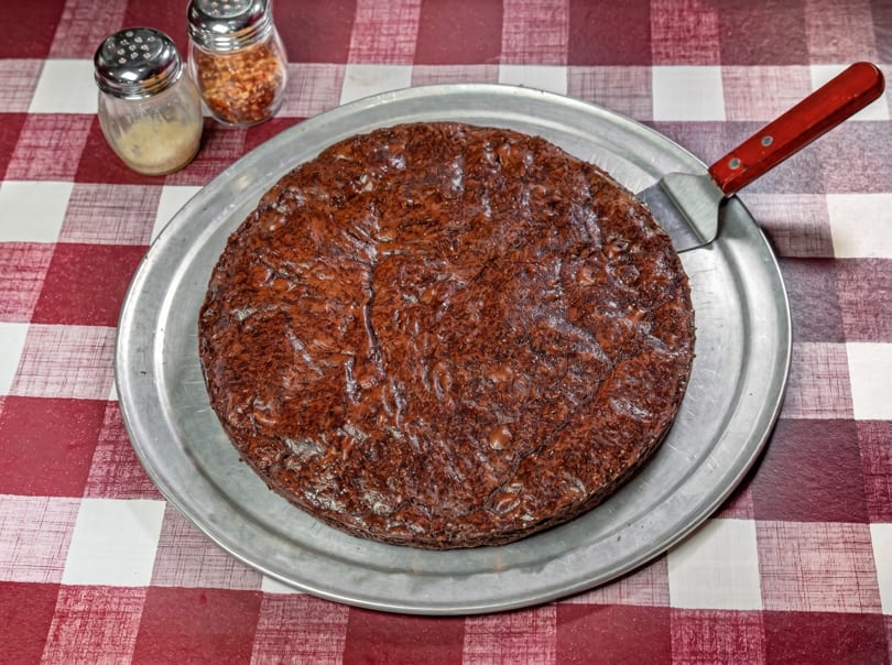 A large, round, dark brown brownie on a metal pizza pan with a red-handled spatula.
