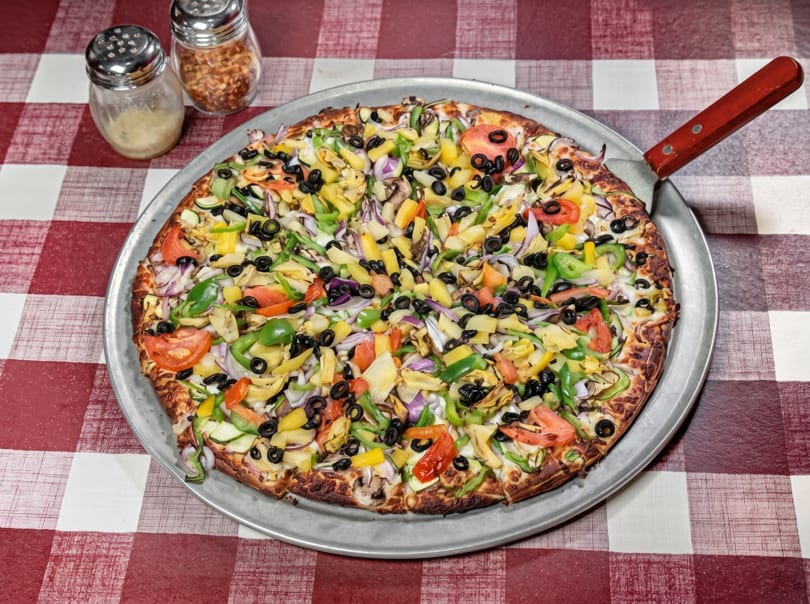 A large, colorful vegetable pizza on a metal pan with a server, on a red and white checkered tablecloth.