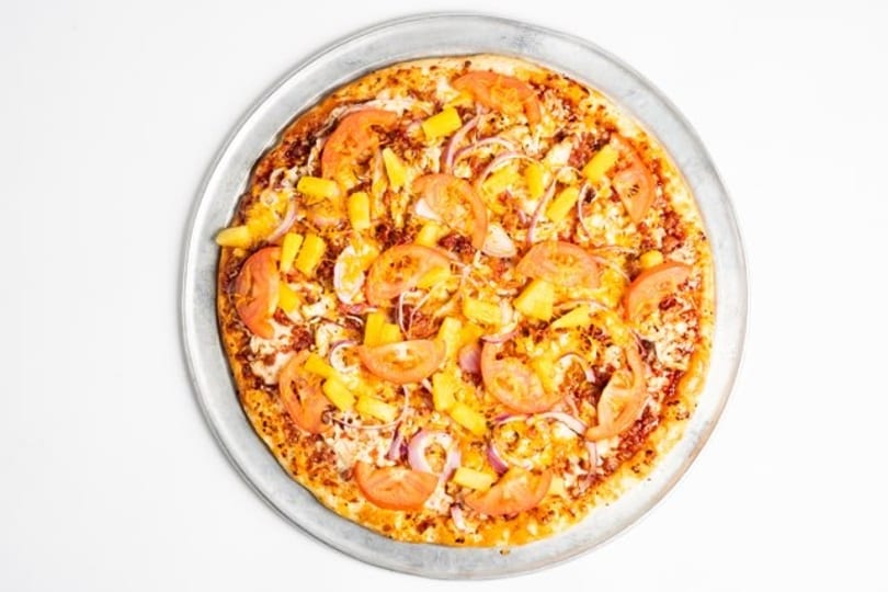 A whole pizza with pineapple, tomatoes, and red onions on a metal pan.