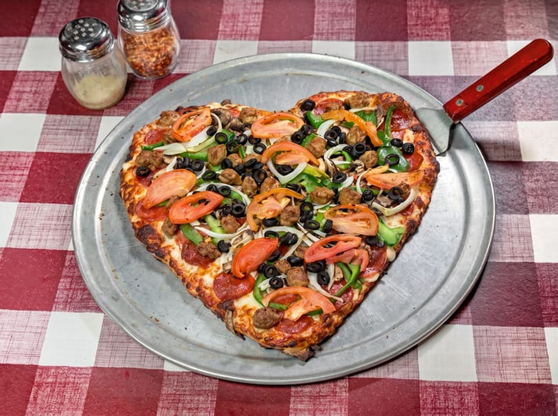 Heart-shaped pizza with pepperoni, sausage, olives, peppers, onions, and tomatoes on a metal tray.