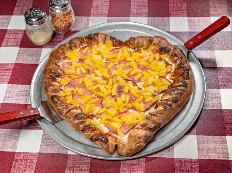 Heart-shaped pizza with ham and pineapple on a metal tray, with shakers on a red and white checkered tablecloth.