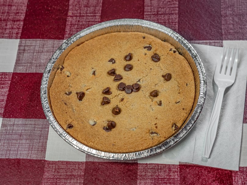 A large chocolate chip cookie in a foil pan with a white plastic fork on a red and white checkered tablecloth.