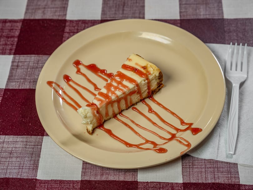A slice of cheesecake drizzled with strawberry sauce on a beige plate with a white fork.